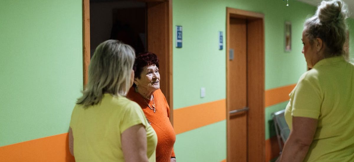 Senior woman engages in conversation with caregivers in a facility corridor.