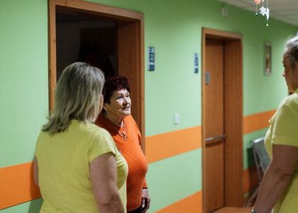 Senior woman engages in conversation with caregivers in a facility corridor.