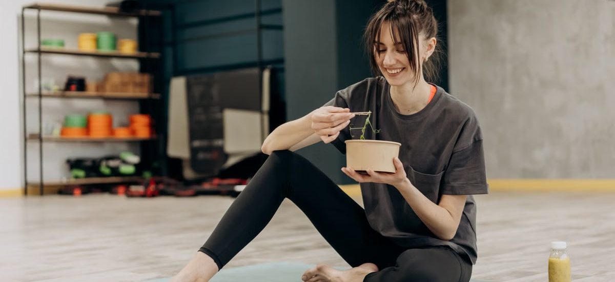 Young woman enjoying a healthy salad after exercising indoors. Promotes a balanced diet and fitness lifestyle.